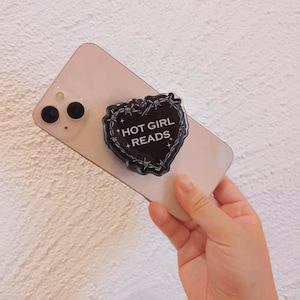 May include: A black heart-shaped phone grip with barbed wire detailing and the text "HOT GIRL READS" in white. The grip is attached to a smartphone.