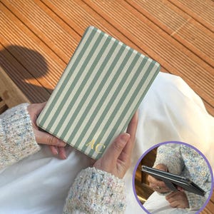 Personalized sage stripe Kindle case held while reading outdoors. Custom initial Kindle cover with retro stripe pattern designed for Kindle Paperwhite and Kindle 2024. Slim protective reader case with comfortable grip for everyday reading.
