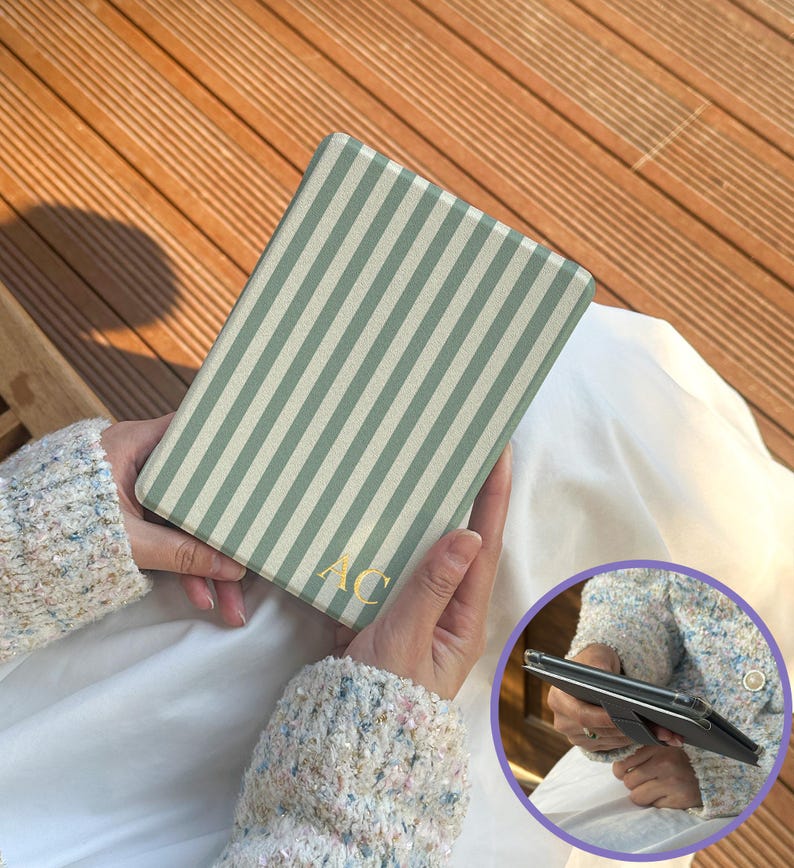 Personalized sage stripe Kindle case held while reading outdoors. Custom initial Kindle cover with retro stripe pattern designed for Kindle Paperwhite and Kindle 2024. Slim protective reader case with comfortable grip for everyday reading.