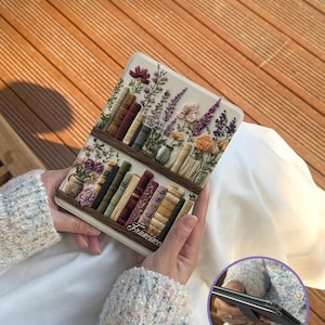 Puede incluir: Una funda para tableta con un diseño bordado de estanterías llenas de libros y flores. La funda tiene un fondo beige, estanterías marrones y la palabra "Francisco" escrita en la estantería inferior.