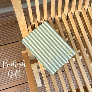 Puede incluir: Un cuaderno rectangular con un patr&oacute;n de rayas verticales en verde salvia y blanco. La cubierta muestra las iniciales AB en color dorado en la esquina inferior derecha. El texto Bookish Gift est&aacute; impreso en blanco a la izquierda.