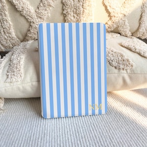Puede incluir: Una funda rectangular para lector electr&oacute;nico con un patr&oacute;n de rayas verticales en azul claro y blanco. La esquina inferior derecha muestra las letras del monograma NM en color dorado con fuente serif. La funda es lisa.