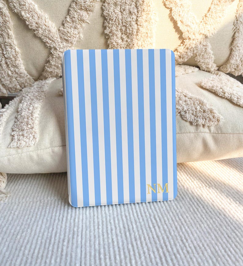 Puede incluir: Una funda rectangular para lector electr&oacute;nico con un patr&oacute;n de rayas verticales en azul claro y blanco. La esquina inferior derecha muestra las letras del monograma NM en color dorado con fuente serif. La funda es lisa.