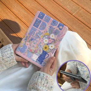 May include: A pink tablet case featuring a floral design with a window view. The artwork includes a blue vase, various flowers, and the text "Slayer Francisco". The case is held open, revealing the tablet's screen.