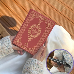 Peut inclure: Un étui rouge en forme de livre avec des motifs floraux et d'oiseaux dorés. L'étui est de forme rectangulaire et est tenu dans les mains. Le mot "Francesca" est imprimé en bas.