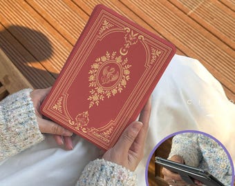 Puede incluir: Una funda roja con forma de libro con diseños florales y de pájaros dorados. La funda tiene forma rectangular y se sostiene con las manos. La palabra "Francesca" está impresa en la parte inferior.