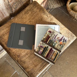 May include: An open e-reader with a decorative cover featuring a bookshelf design with flowers and books. The e-reader is on a wooden surface, next to a gray case.