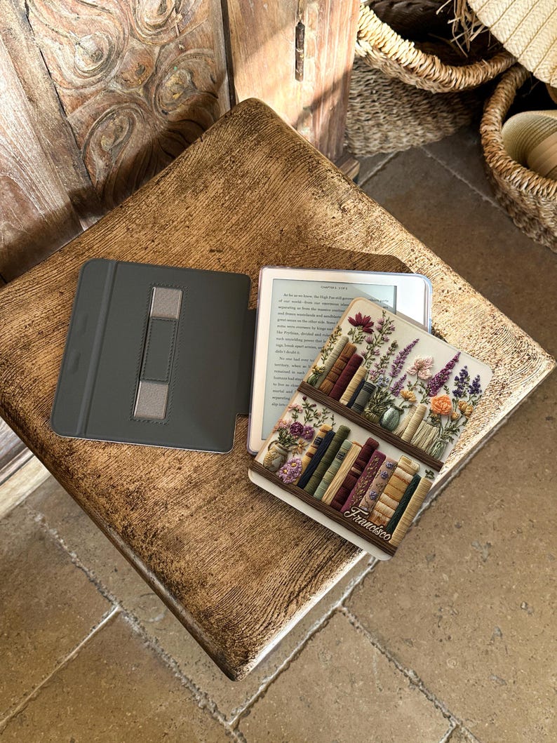 May include: An open e-reader with a decorative cover featuring a bookshelf design with flowers and books. The e-reader is on a wooden surface, next to a gray case.
