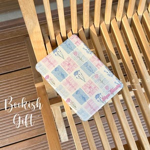 May include: A rectangular book cover with a floral pattern in pink and blue squares, resting on a wooden chair. The text "Bookish Gift" is written in cursive on the left side of the image.