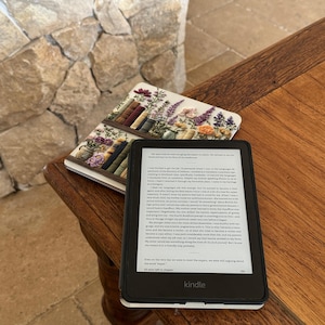 May include: A black Kindle e-reader with a white border displays text on its screen, resting on a wooden table. A floral-themed notebook with a bookshelf design lies beside it. The background features a stone wall.