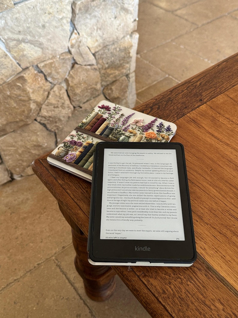 May include: A black Kindle e-reader with a white border displays text on its screen, resting on a wooden table. A floral-themed notebook with a bookshelf design lies beside it. The background features a stone wall.