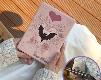 May include: A pink tablet case featuring a black bat silhouette within a heart-shaped floral design, a pink heart, and the text "Slayer Frances". The case is held in a person's hands.