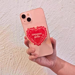 May include: A pink smartphone with a red heart-shaped phone grip. The grip features the words "romance girlie" in white script, surrounded by a barbed wire design. The phone is held in a hand against a textured white background.