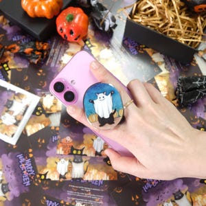 Può includere: Uno smartphone rosa con un supporto per telefono a tema Halloween con un fantasma dei cartoni animati e zucche. Lo sfondo include biglietti di Halloween, zucche decorative e una scatola nera con paglia.
