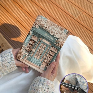 May include: A tablet case featuring a bookstore illustration with the text "Book Store". The case has a teal door and window design, surrounded by white flowers. The case is held by a person wearing a light-colored sweater.