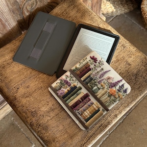 May include: An open e-reader with a dark gray cover and a decorative book-themed coaster. The coaster features a floral design with a stack of books and the name "Francisco". The e-reader is on a wooden surface.