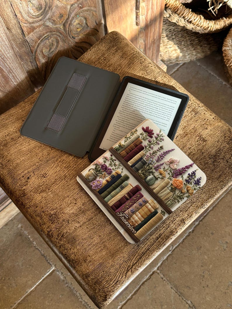 May include: An open e-reader with a dark gray cover and a decorative book-themed coaster. The coaster features a floral design with a stack of books and the name "Francisco". The e-reader is on a wooden surface.