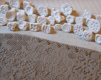 May include: A collection of white ceramic stamps with various designs, including geometric patterns, flowers, and swirls. The stamps are arranged on a white surface, and some have been used to create impressions in a brown clay surface.