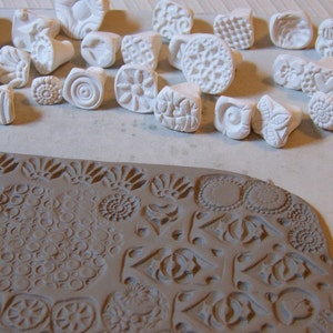 Pu&ograve; includere: Una collezione di timbri in ceramica bianca con vari disegni, tra cui motivi geometrici, fiori e volute. I timbri sono disposti su una superficie bianca, e una superficie in argilla marrone mostra le impressioni fatte dai timbri.