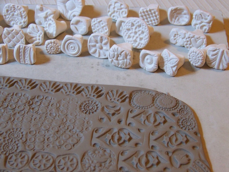 Pu&ograve; includere: Una collezione di timbri in ceramica bianca con vari disegni, tra cui motivi geometrici, fiori e volute. I timbri sono disposti su una superficie bianca, e una superficie in argilla marrone mostra le impressioni fatte dai timbri.
