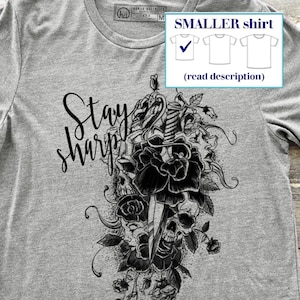 May include: Gray t-shirt with a crew neck and short sleeves. The shirt features a black graphic of a sword, roses, and skulls, with the words "Stay Sharp" in a script font. A smaller shirt size is indicated.