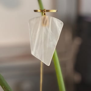 May include: A delicate, pearlescent white pendant with a gold-colored ring and chain. The pendant has a unique, asymmetrical shape and hangs from a thin, gold-colored chain. The background features a green stem.