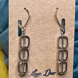 May include: A pair of dangle earrings featuring three stacked, clear, square-shaped beads with rounded corners, suspended from gold-tone metal ear wires. The earrings are displayed on a rectangular tan card labeled Magic Deer Designs.