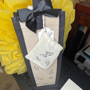 May include: A beige burlap gift bag with black trim and a black satin ribbon bow. The bag has a small white fabric pouch with a black olive branch design inside. The bag has a white tag with the word "Jo." written on it.