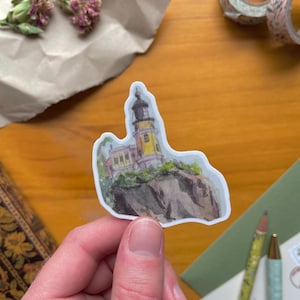 May include: A sticker featuring a watercolor illustration of a yellow lighthouse atop a rocky cliff. The lighthouse has a black top and is surrounded by green foliage. The sticker is held by a hand against a wooden background.