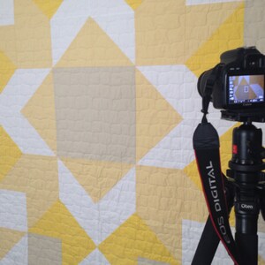 May include: A black DSLR camera mounted on a tripod with a black and red strap. The camera is pointed at a white, yellow, and beige quilt with a geometric pattern.