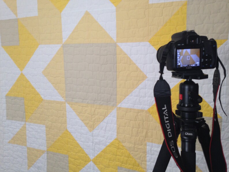 May include: A black DSLR camera mounted on a tripod with a black and red strap. The camera is pointed at a white, yellow, and beige quilt with a geometric pattern.
