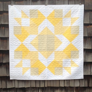 May include: A white quilt with a geometric pattern in shades of yellow and beige. The quilt is made up of squares and triangles, and the pattern is symmetrical.