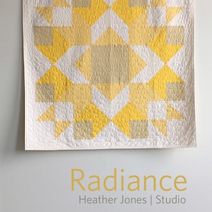 May include: A white quilt with a geometric pattern of yellow and beige squares and triangles. The quilt is hanging on a white wall with the text "Radiance Heather Jones | Studio" below it.