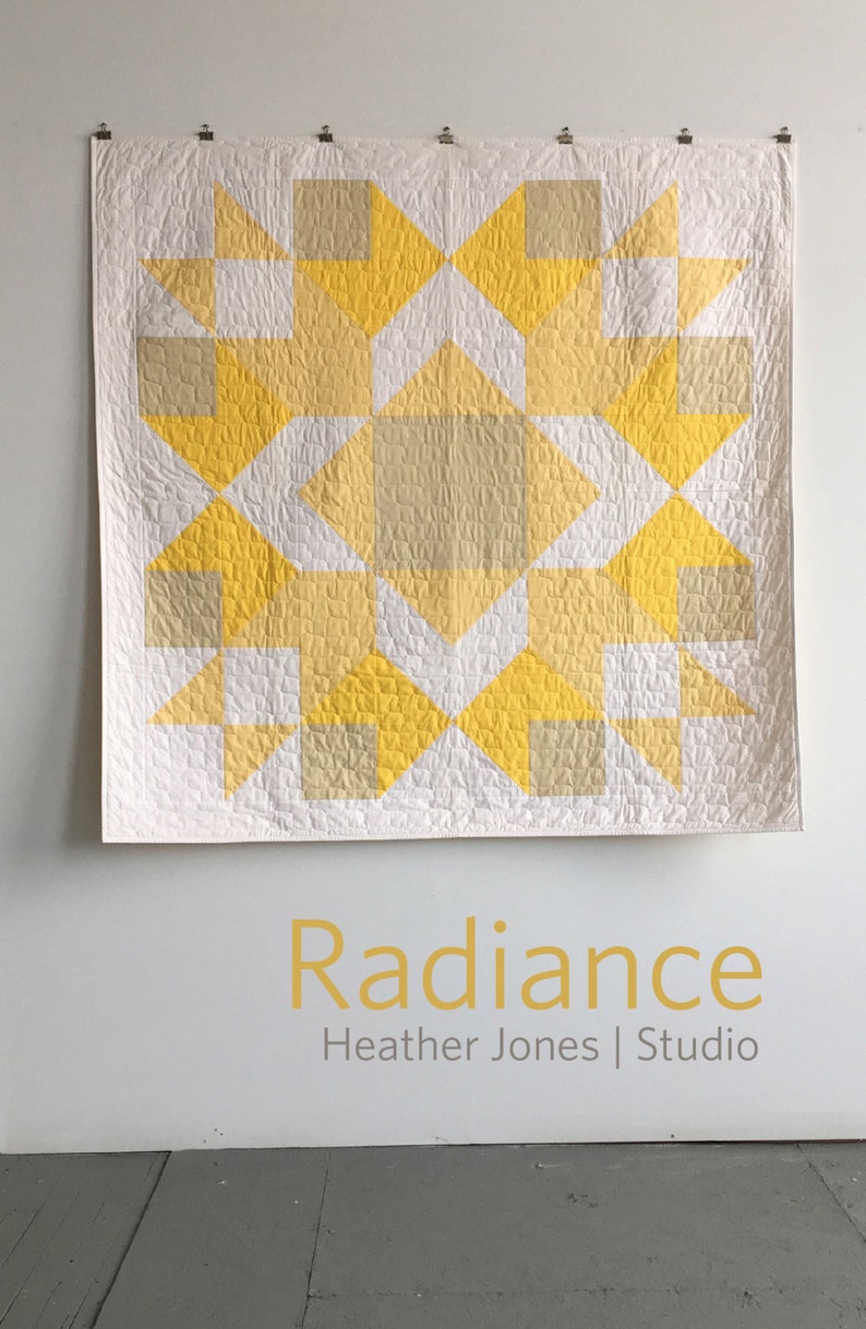 May include: A white quilt with a geometric pattern of yellow and beige squares and triangles. The quilt is hanging on a white wall with the text "Radiance Heather Jones | Studio" below it.