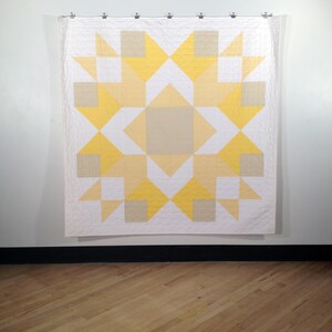 May include: A white quilt with a yellow and beige geometric pattern. The quilt is hanging on a white wall.