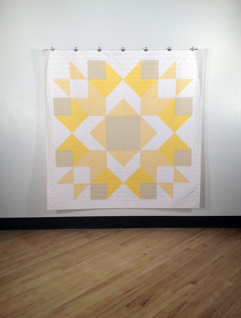 May include: A white quilt with a yellow and beige geometric pattern. The quilt is hanging on a white wall.