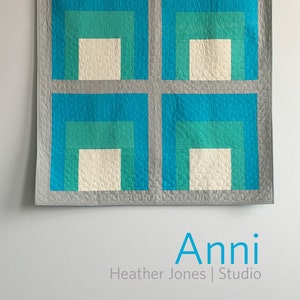 May include: A gray and white quilt with four squares, each containing a concentric pattern of turquoise, teal, and white squares. The quilt is hanging on a white wall with the text "Anni Heather Jones | Studio" below it.