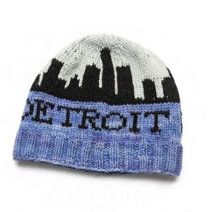 May include: A knit beanie featuring a light gray top section with a black skyline silhouette. The lower section is a variegated blue with the word DETROIT knitted in black block letters across the front. The hat has a ribbed cuff edge.