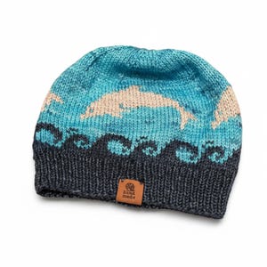 May include: A knitted beanie hat with a dolphin design. The hat features a blue and teal ocean scene with a beige dolphin leaping through the waves. The bottom of the hat is dark gray, and a small brown tag reads "hand made".