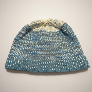 May include: A knitted beanie hat in shades of blue, white, and gray. The hat features a ribbed brim and a textured pattern throughout the body. The top of the hat is a lighter color, transitioning from the darker blue base.