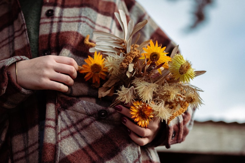 May include: A person wearing a brown and white plaid shirt is holding a bouquet of dried flowers. The flowers are a mix of yellow, brown, and white.