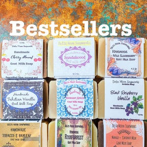May include: Nine rectangular bars of handmade goat milk soap arranged in a grid, each wrapped in a paper label featuring unique designs and scents including Cherry Almond, Sandalwood, Wild Elderberry, Tahitian Vanilla, Hawaiian Ginger, and Mango.