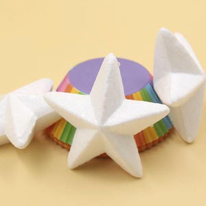 May include: Three white foam star shapes on top of a rainbow cupcake liner. The stars are 3-dimensional and have a slightly textured surface.