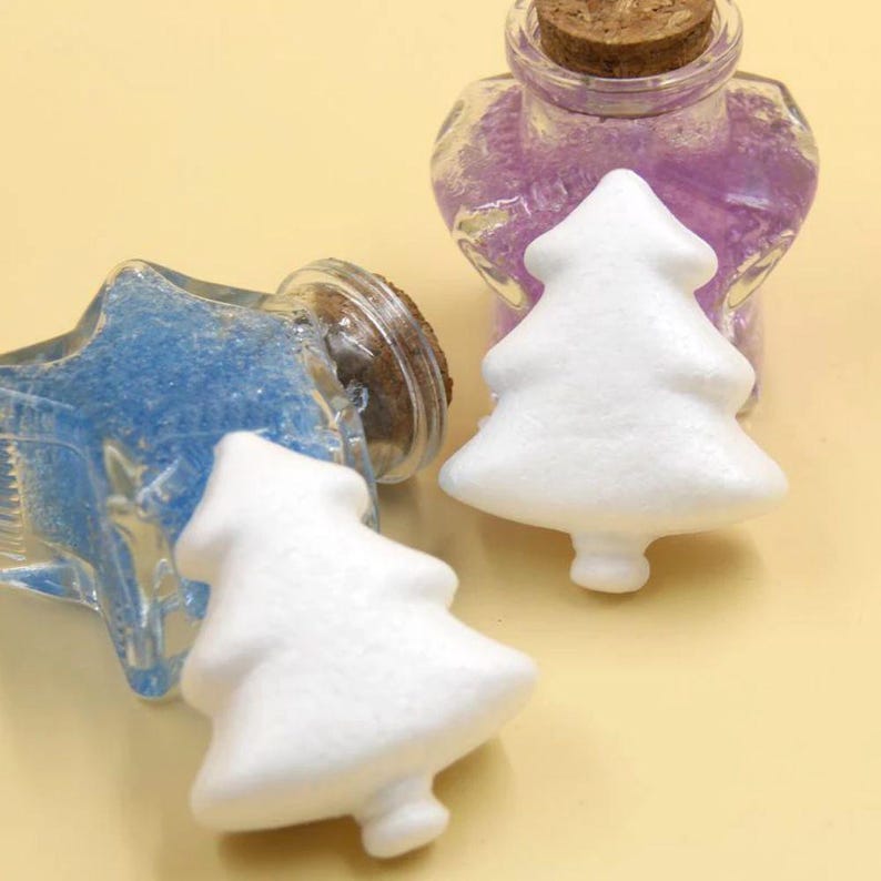 May include: Two glass bottles filled with blue and purple glitter, each with a white foam Christmas tree ornament on top. The bottles have cork stoppers.