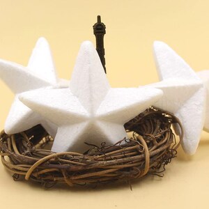 May include: Three white foam star ornaments arranged in a wreath of brown twigs. The stars are arranged with points facing outward, creating a circular shape.
