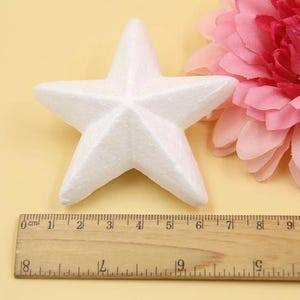 May include: A white foam star measuring 6 inches across. The star is sitting on a yellow surface next to a pink flower and a wooden ruler.