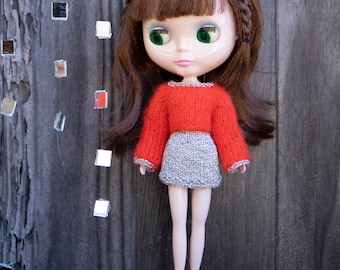 May include: A doll with brown hair and green eyes wearing a red sweater and a gray skirt. The doll has a braided hairstyle with small flower hair accessories and is wearing lavender shoes. The background is a weathered wooden surface.