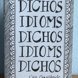 May include: A small, hand-drawn book featuring the words "DICHOS IDIOMS" repeated in bold, black lettering. The title is framed by a decorative border and the author's name, "Liz Comstock," is at the bottom. The background is light blue.