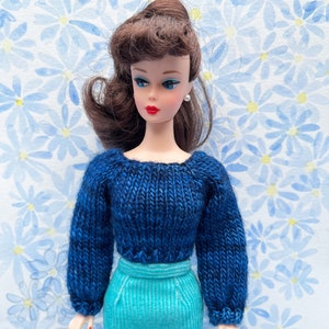 May include: A vintage doll with brown hair wearing a blue knitted sweater and a turquoise skirt. The doll is standing in front of a blue and white floral background.