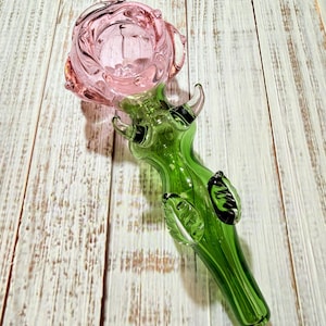 May include: A glass pipe shaped like a rose flower with a translucent pink bowl and a green stem. The stem features small green glass leaves and thorns extending from the sides, leading to a tapered mouthpiece at the bottom of the glass object.